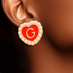 Heart-shaped earring with red center and letter 'G' worn on an ear against a brown background