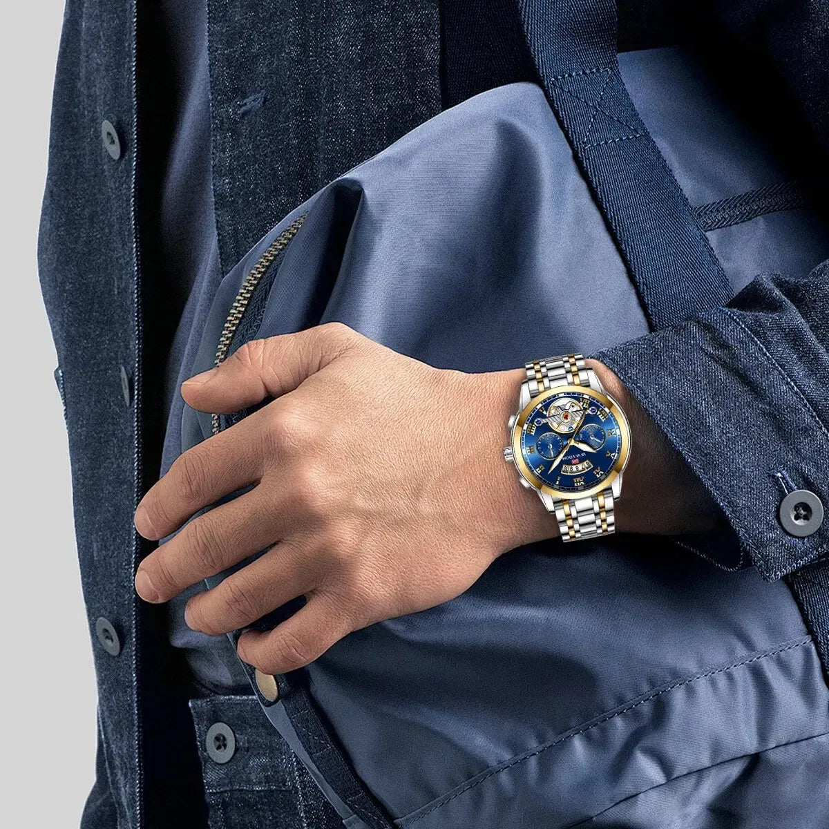 Hand wearing a gold and blue watch with a blue jacket on a gray background